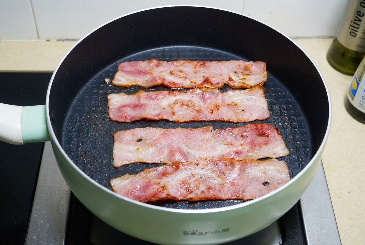 Quatre tranches de bacon grillant dans une poêle antiadhésive noire jusquà ce que le gras bouillonne et que la viande soit dorée et croustillante.