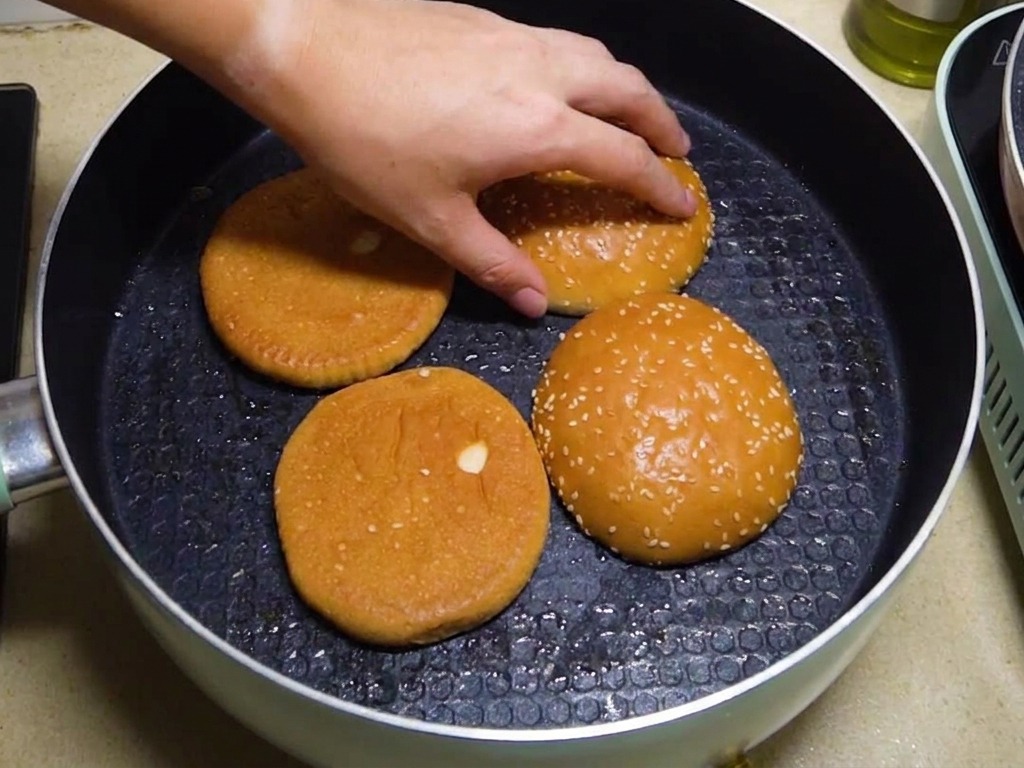 Quatre moitiés de pain à burger toastées face vers le bas dans une poêle, avec une main ajustant leur position.