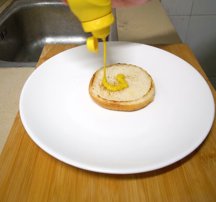 Une main versant de la moutarde jaune vif depuis une bouteille en plastique compressible sur un pain à burger grillé posé dans une assiette blanche.
