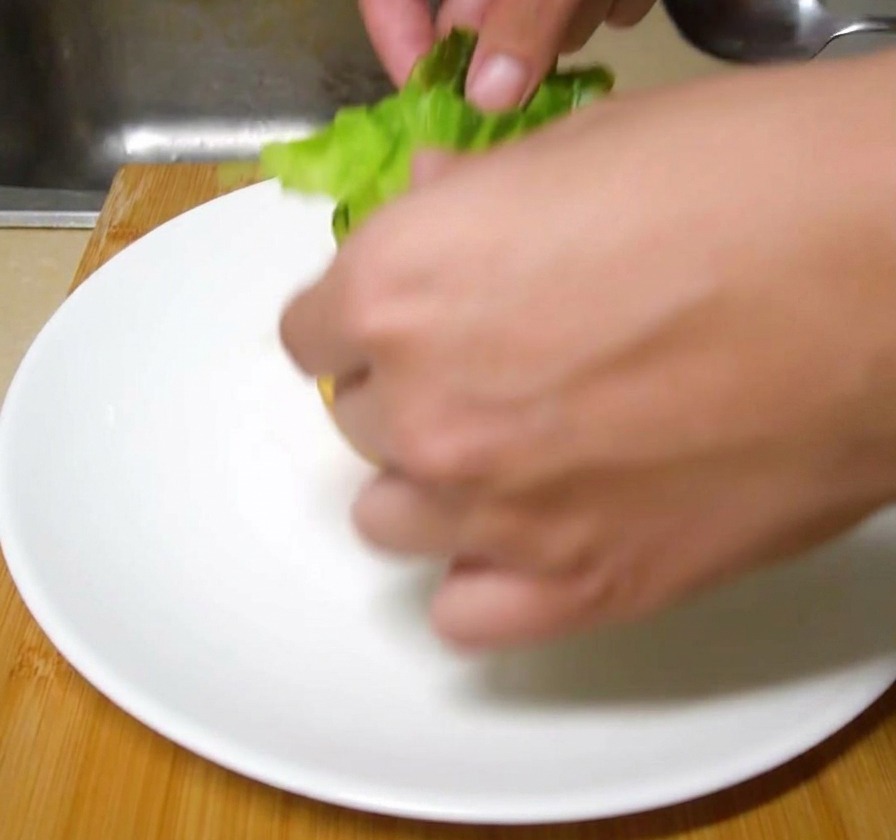 Des feuilles de laitue verte fraîche déposées à la main sur un pain à burger grillé dans une assiette en céramique blanche.