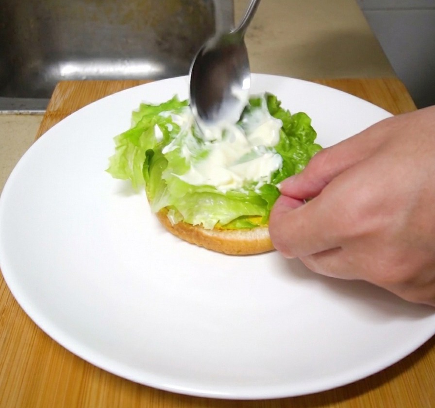 Une cuillère en métal étalant une noix de mayonnaise blanche sur des feuilles de laitue verte fraîche dans un burger en cours dassemblage.