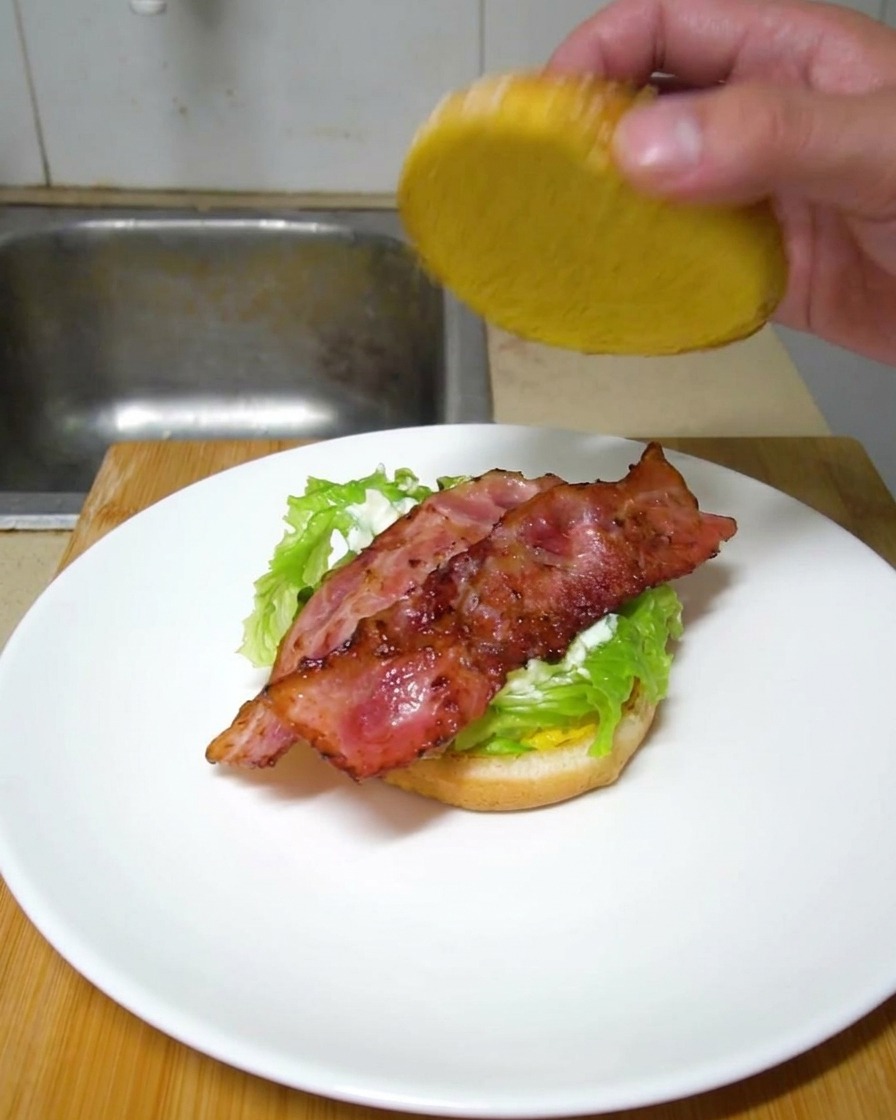 Une main déposant le pain supérieur sur un burger composé de laitue et de tranches de bacon croustillantes.