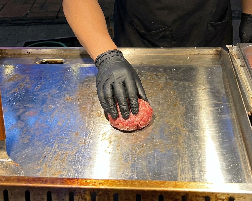 Une main gantée de noir utilisant une presse à viande en métal pour écraser une boule de bœuf haché cru sur une plaque chaude.