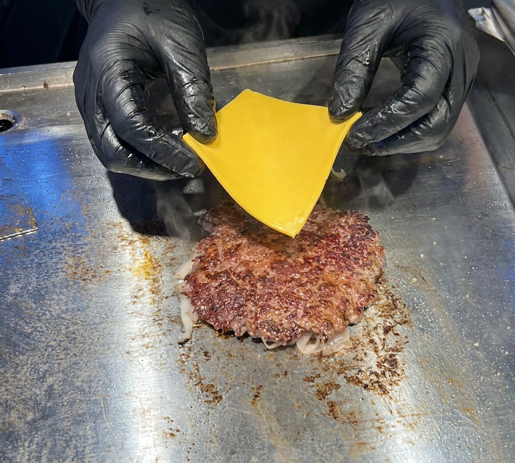 Gros plan de mains gantées de noir déposant délicatement une tranche de cheddar jaune sur un steak de bœuf grésillant.