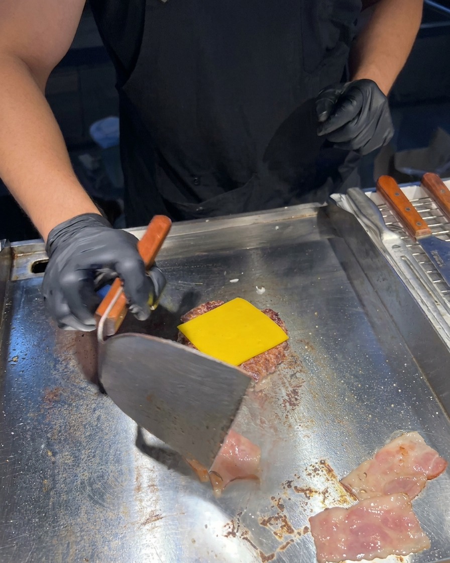 Une spatule en métal retournant des tranches de bacon grésillantes à côté du steak nappé de fromage.