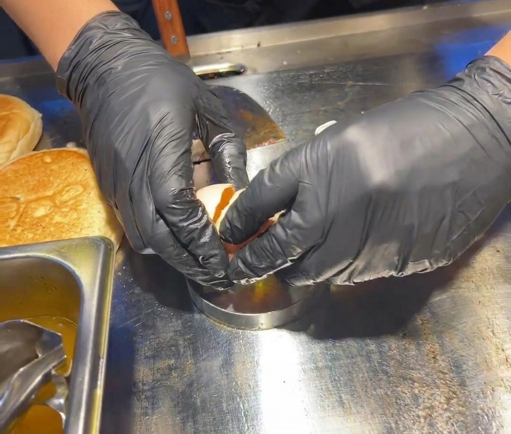 Des mains gantées de noir cassant un œuf frais dans un cercle en métal posé sur la plaque de cuisson chaude.
