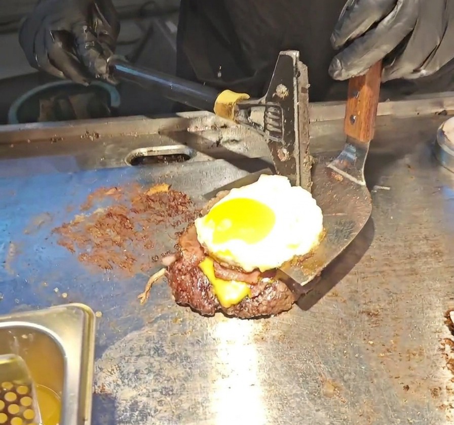 Une large spatule en métal soulevant la superposition du steak, du fromage fondu, du bacon et de lœuf au plat coulant.