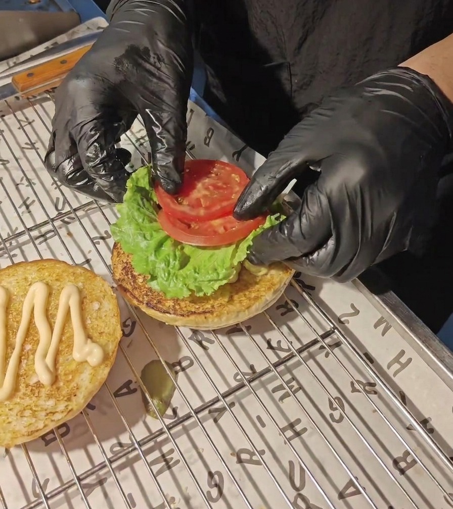 Des mains gantées de noir déposant des tranches de tomate fraîche sur une feuille de laitue verte.