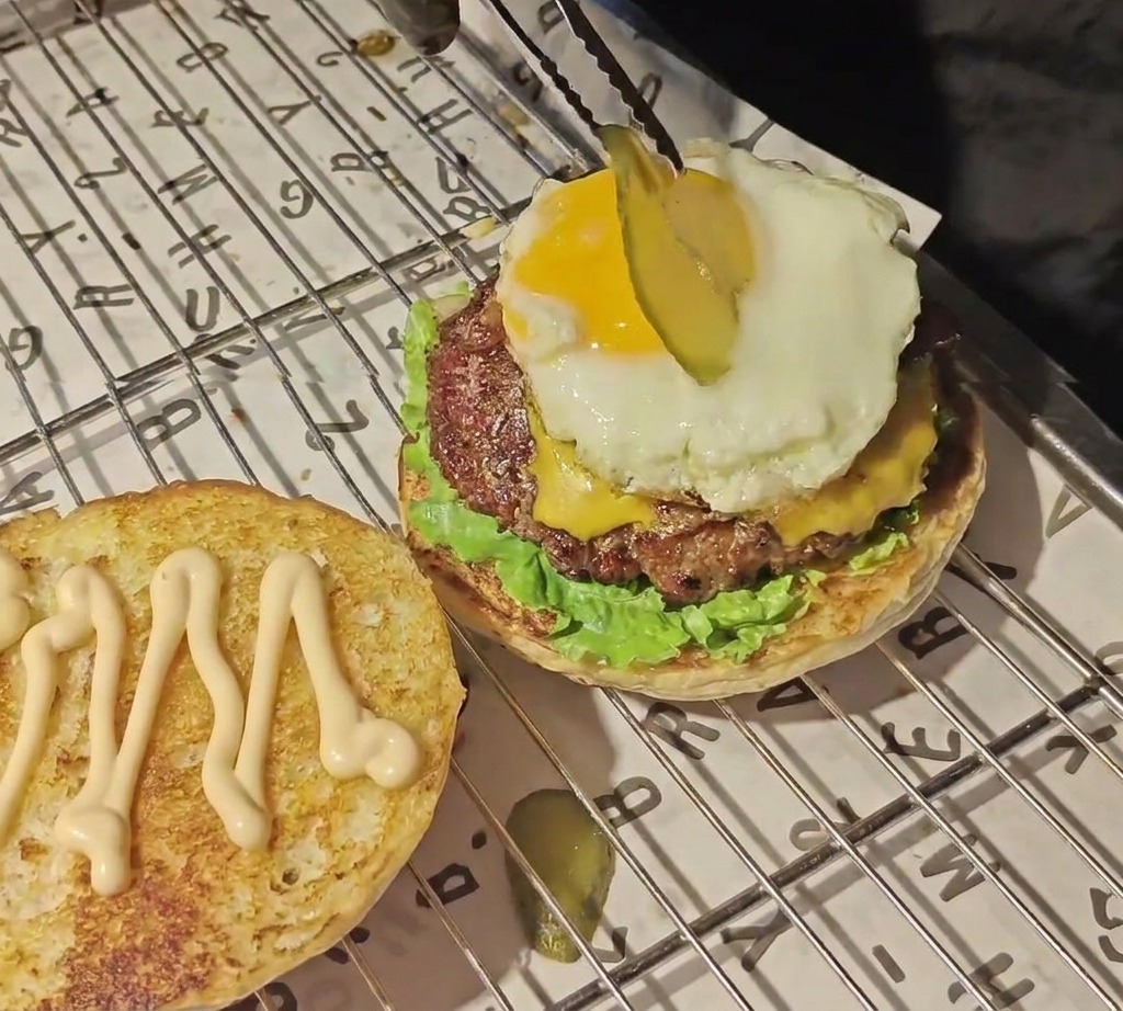 Une pince déposant une fine rondelle de cornichon sur un jaune dœuf éclatant au sommet du burger.