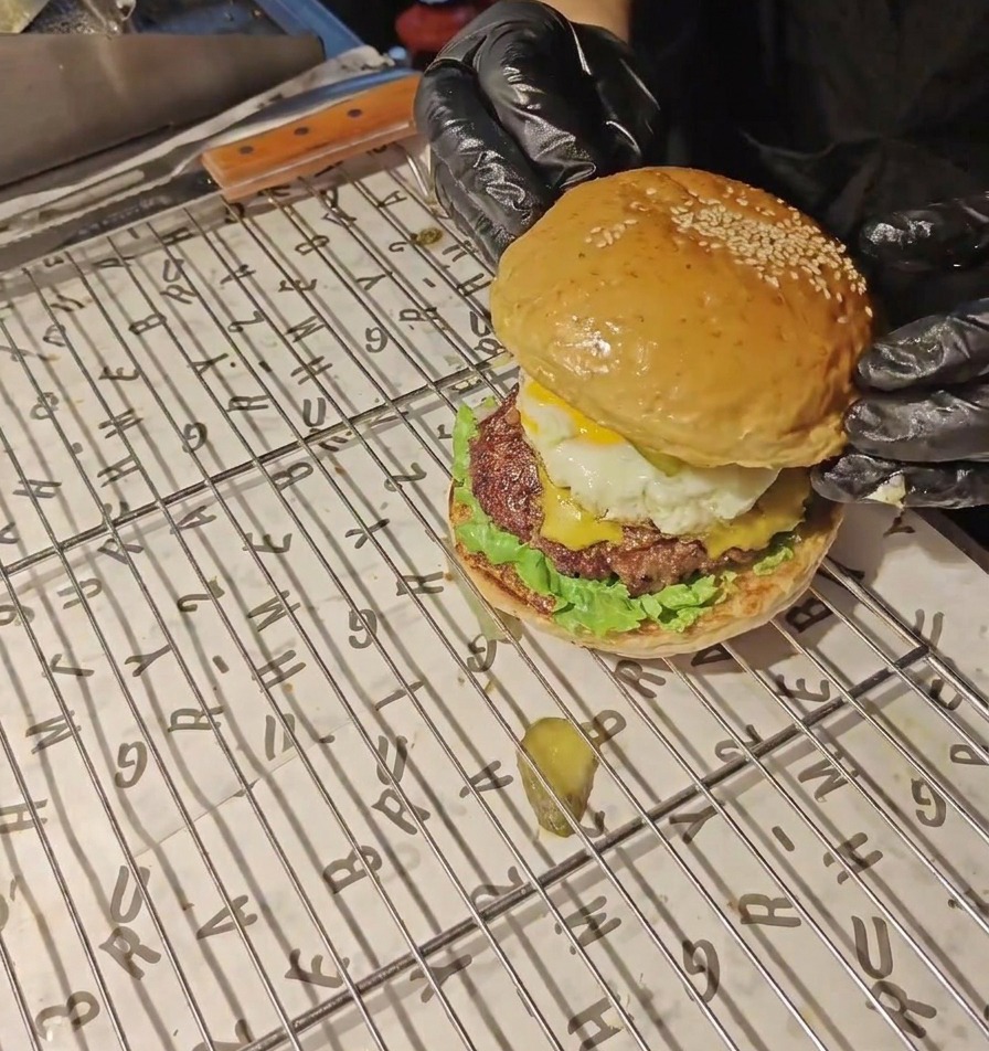 Des mains gantées de noir posant le pain supérieur bien doré sur le burger entièrement assemblé.