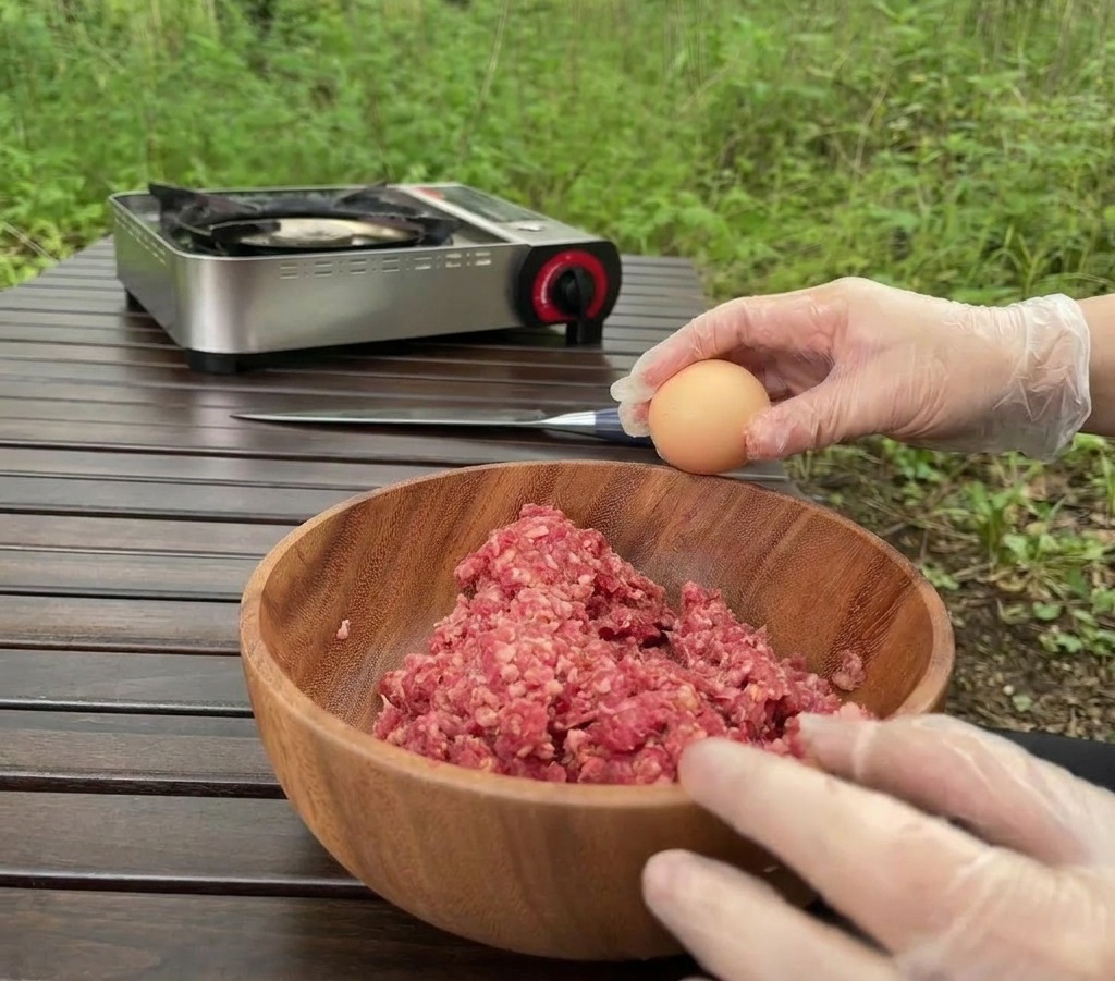 Une personne portant des gants transparents tenant un œuf brun au-dessus dun bol en bois rempli de bœuf haché cru, sur une table en bois à lextérieur.