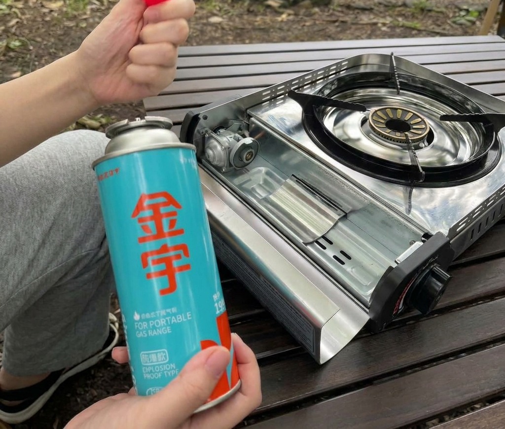 Une personne tenant une cartouche de gaz butane bleue et son capuchon rouge à côté du compartiment ouvert dun réchaud portable.