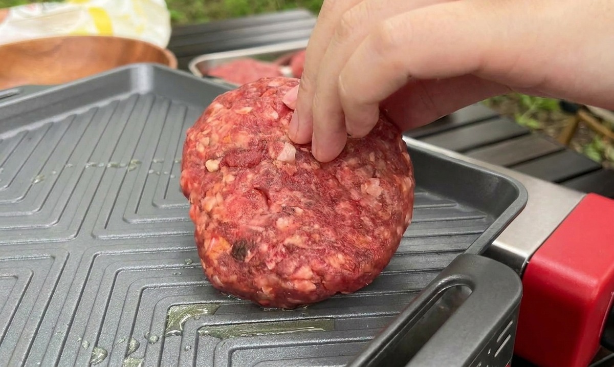 Une main déposant un steak haché cru et épais sur une poêle-gril carrée et chaude lors dune séance de cuisine en plein air.