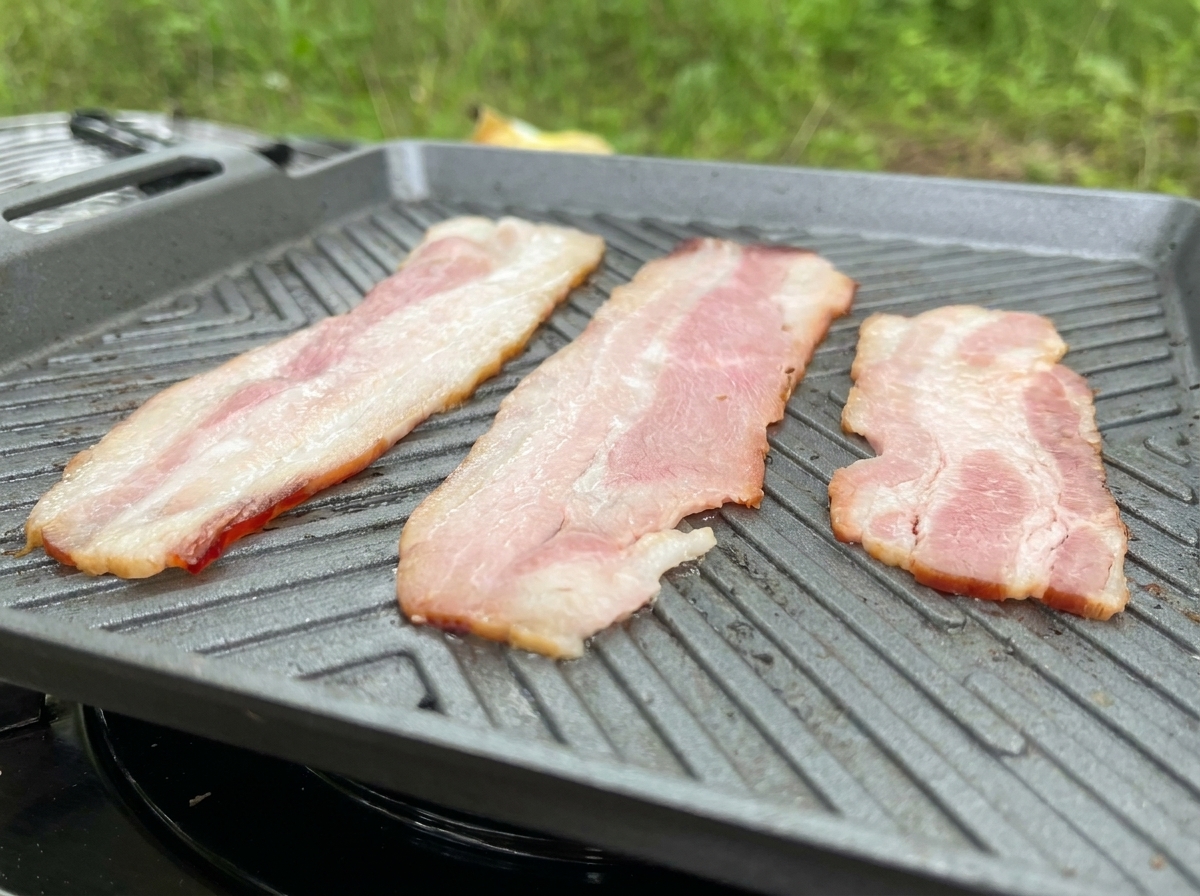 Trois longues tranches de bacon en train de frire sur une plancha grise, avec le gras qui fond.