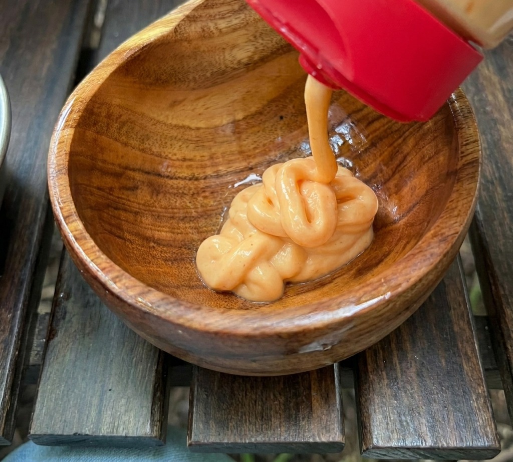 Une sauce orange onctueuse est pressée depuis un flacon à bouchon rouge dans un petit bol en bois contenant dautres ingrédients pour la sauce.