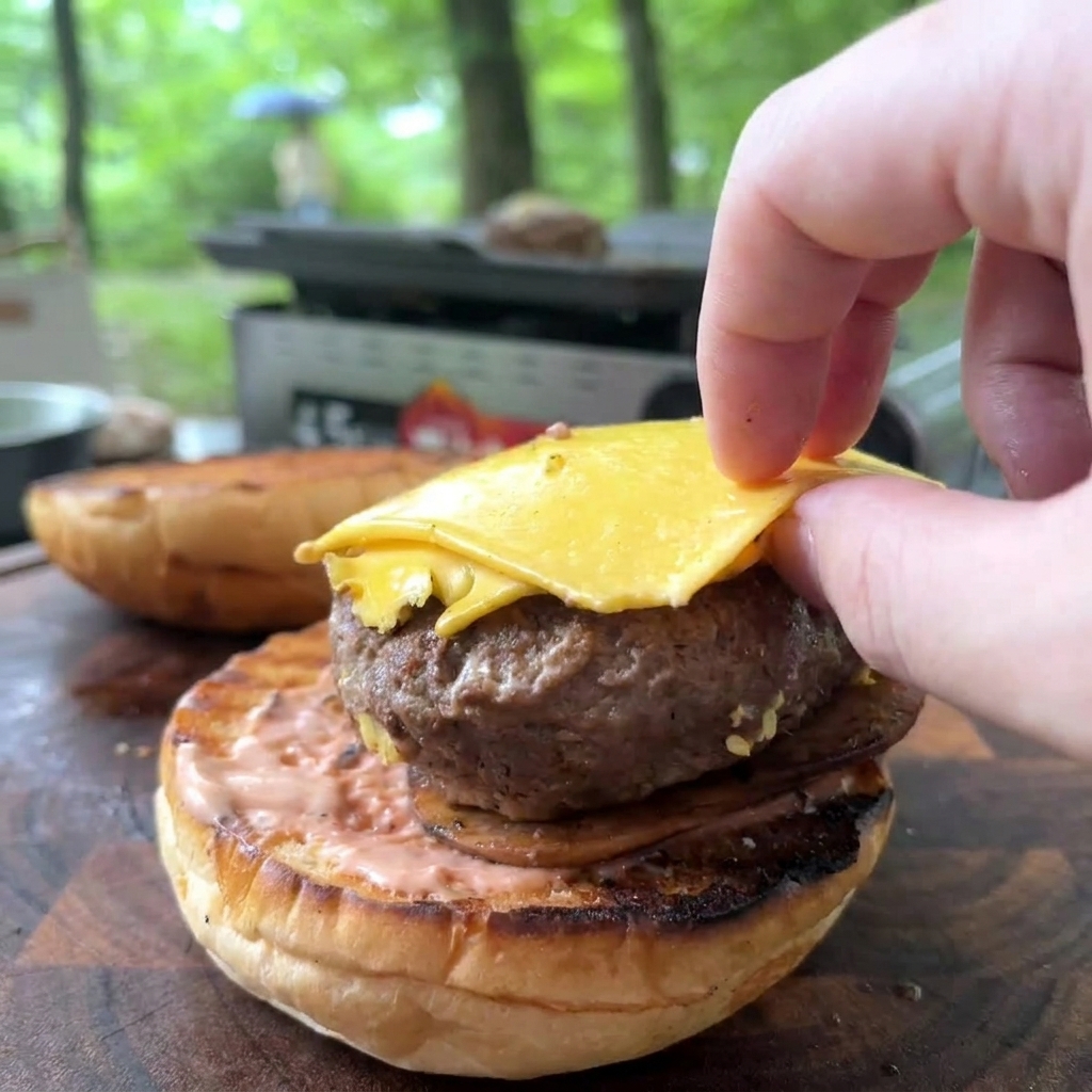 Une main plaçant un steak de bœuf cuit garni de fromage jaune fondu sur un pain toasté tartiné de sauce rose.