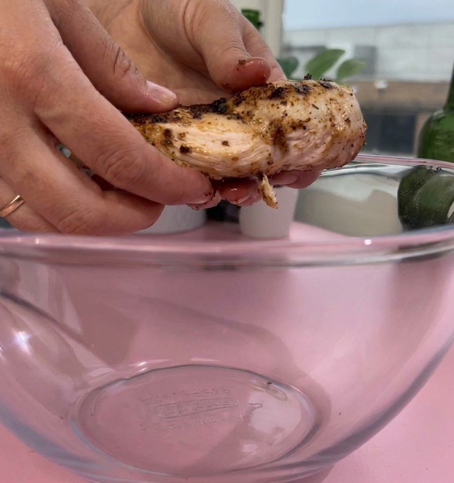 Des mains plaçant un morceau de blanc de poulet cuit et assaisonné dans un grand saladier en verre transparent sur une surface rose.