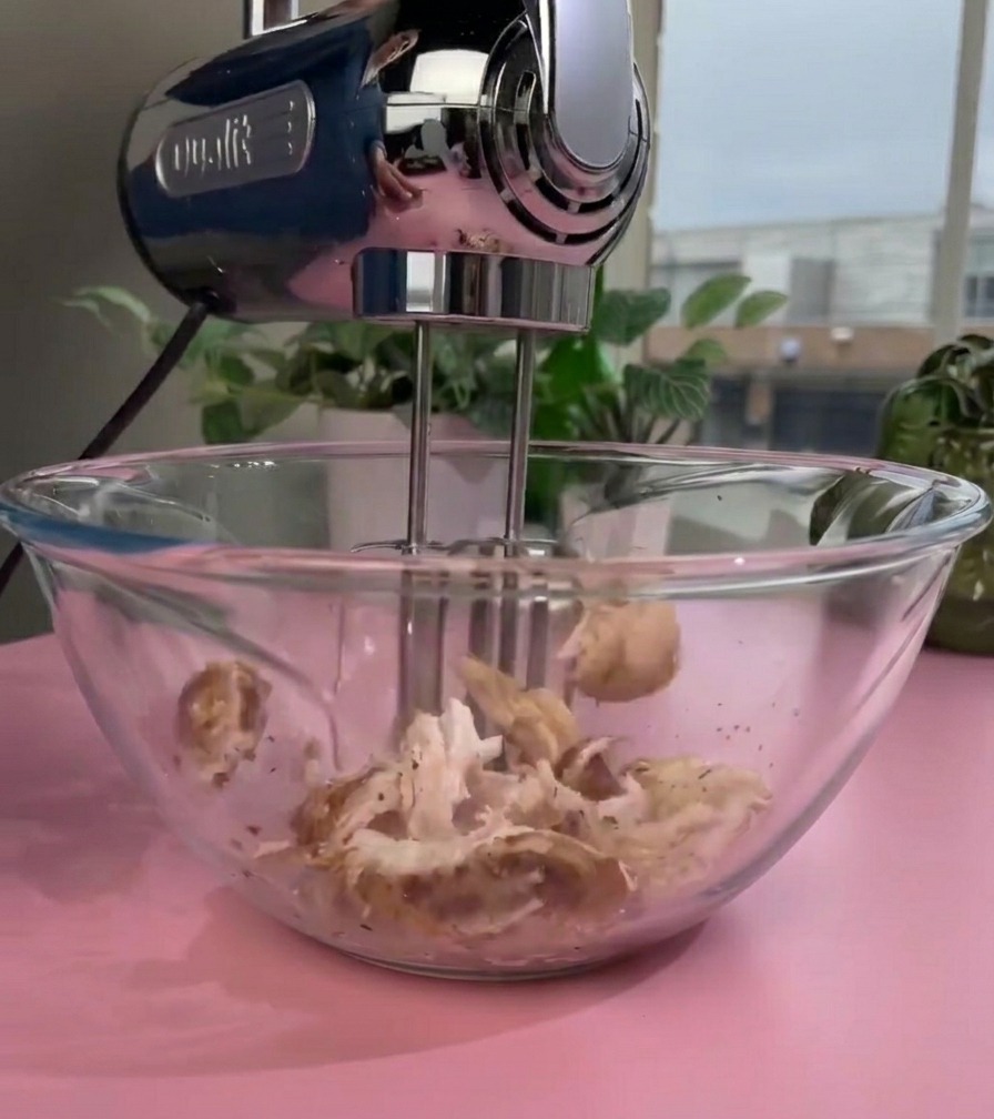 Un batteur électrique doté de fouets rotatifs effilochant du poulet cuit à lintérieur dun saladier en verre pour obtenir une texture fine.