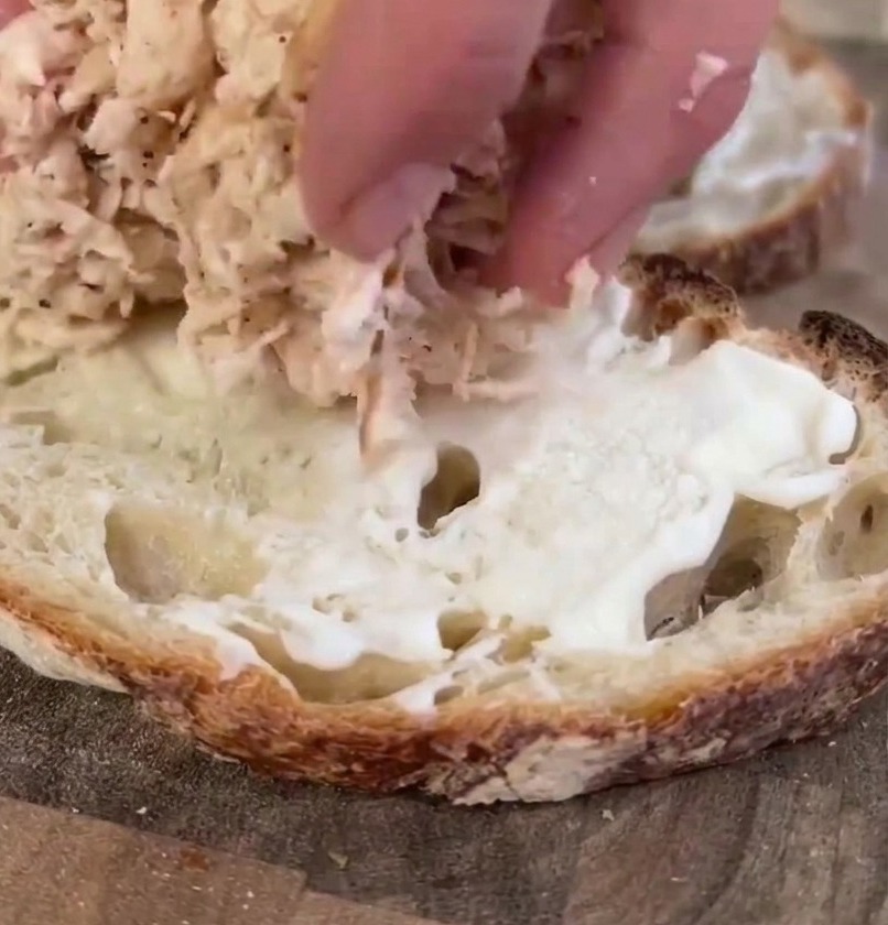 La main dune personne déposant un dôme de poulet cuit effiloché sur une tranche de pain au levain garnie de mayonnaise.