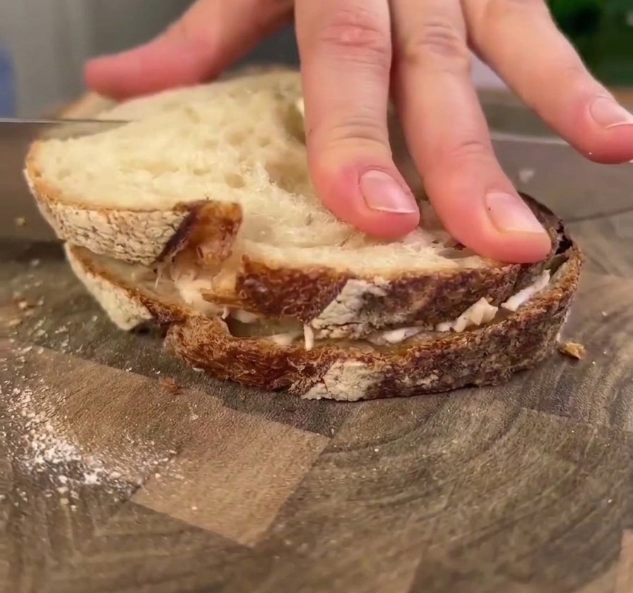 Une main pressant fermement un grand sandwich au poulet et pain au levain sur une planche en bois tandis quun couteau sapprête à le couper.