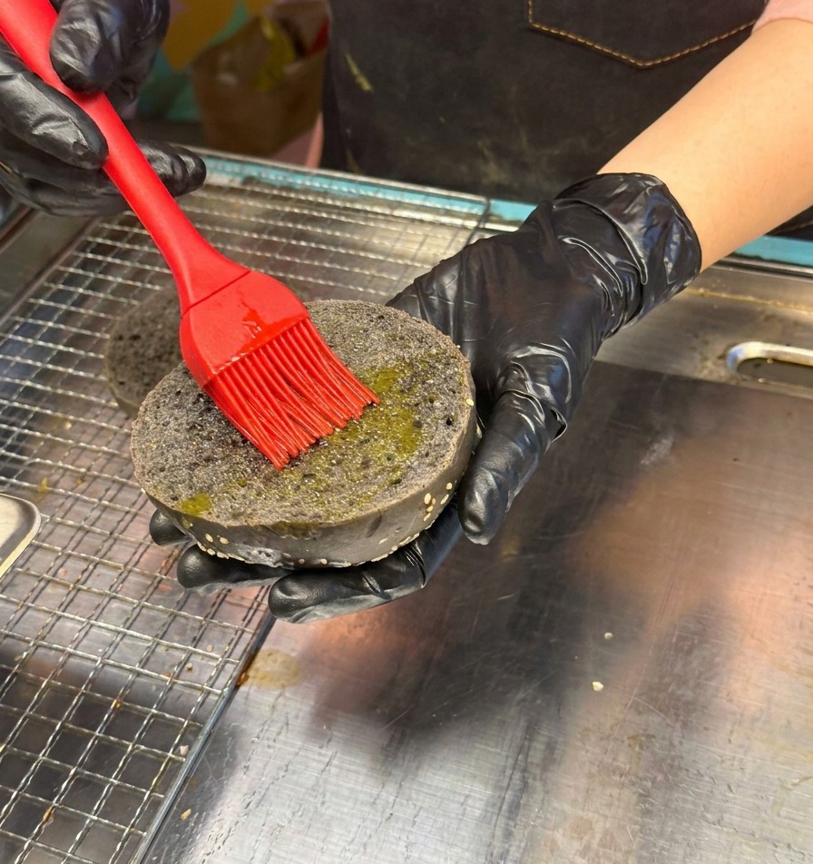 Une main gantée de noir utilisant un pinceau en silicone rouge pour huiler lintérieur dun pain à burger noir aux graines de sésame.
