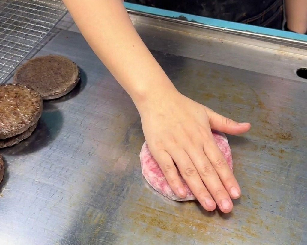 Une main déposant un steak haché de bœuf cru sur une plancha en inox propre, à côté de moitiés de pain toastées.