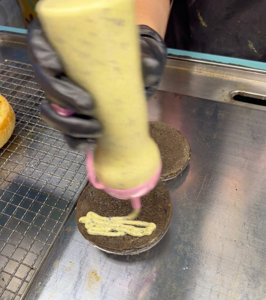 Un flacon à embout répartissant une sauce crémeuse et mouchetée à la truffe en zigzag sur un pain à burger noir.