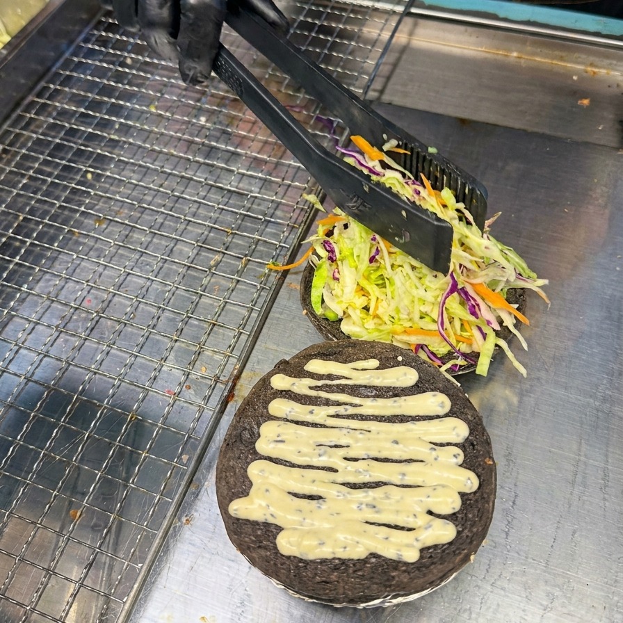 Une personne utilisant une pince pour placer délicatement une tranche de tomate rouge vif sur un lit de crudités dans un pain à burger noir au charbon.