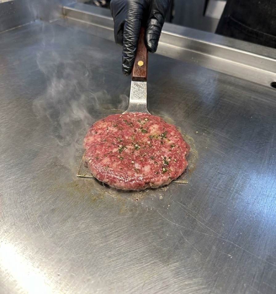 Une spatule en métal pressant un steak de bœuf sur une plancha en inox dégageant de la vapeur.