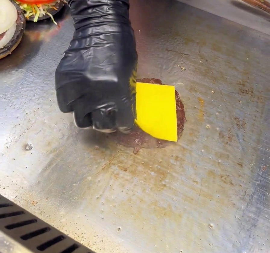 Une main gantée de noir déposant une tranche de fromage jaune sur un steak de burger saisi sur une plaque de cuisson.