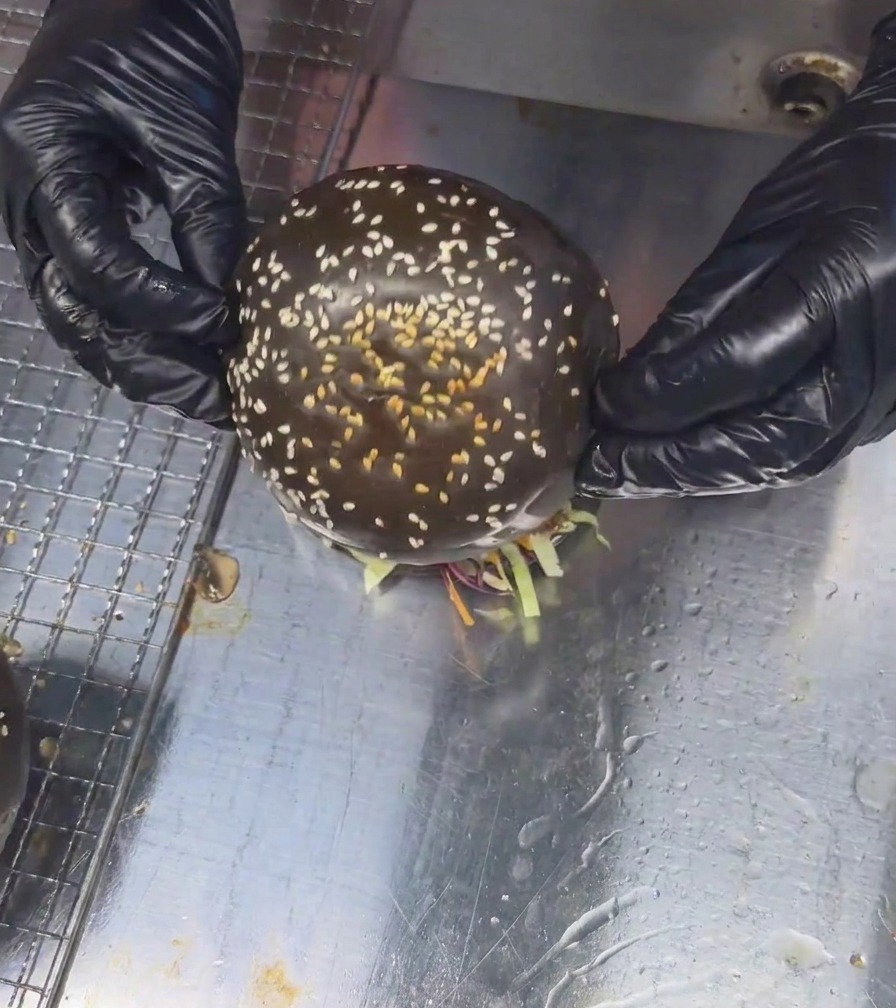 Deux mains gantées plaçant un pain aux graines de sésame noir sur un grand burger au bœuf et aux champignons entièrement monté.
