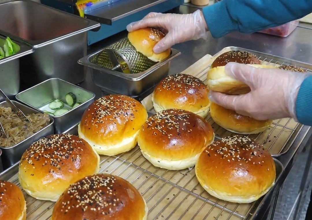 Mains gantées faisant rouler des pains à burger sur un rouleau à beurre rotatif en métal.