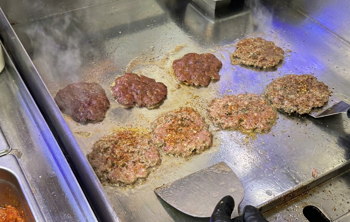 Steaks de bœuf écrasés et saisis sur une plancha en inox à laide dune spatule métallique.