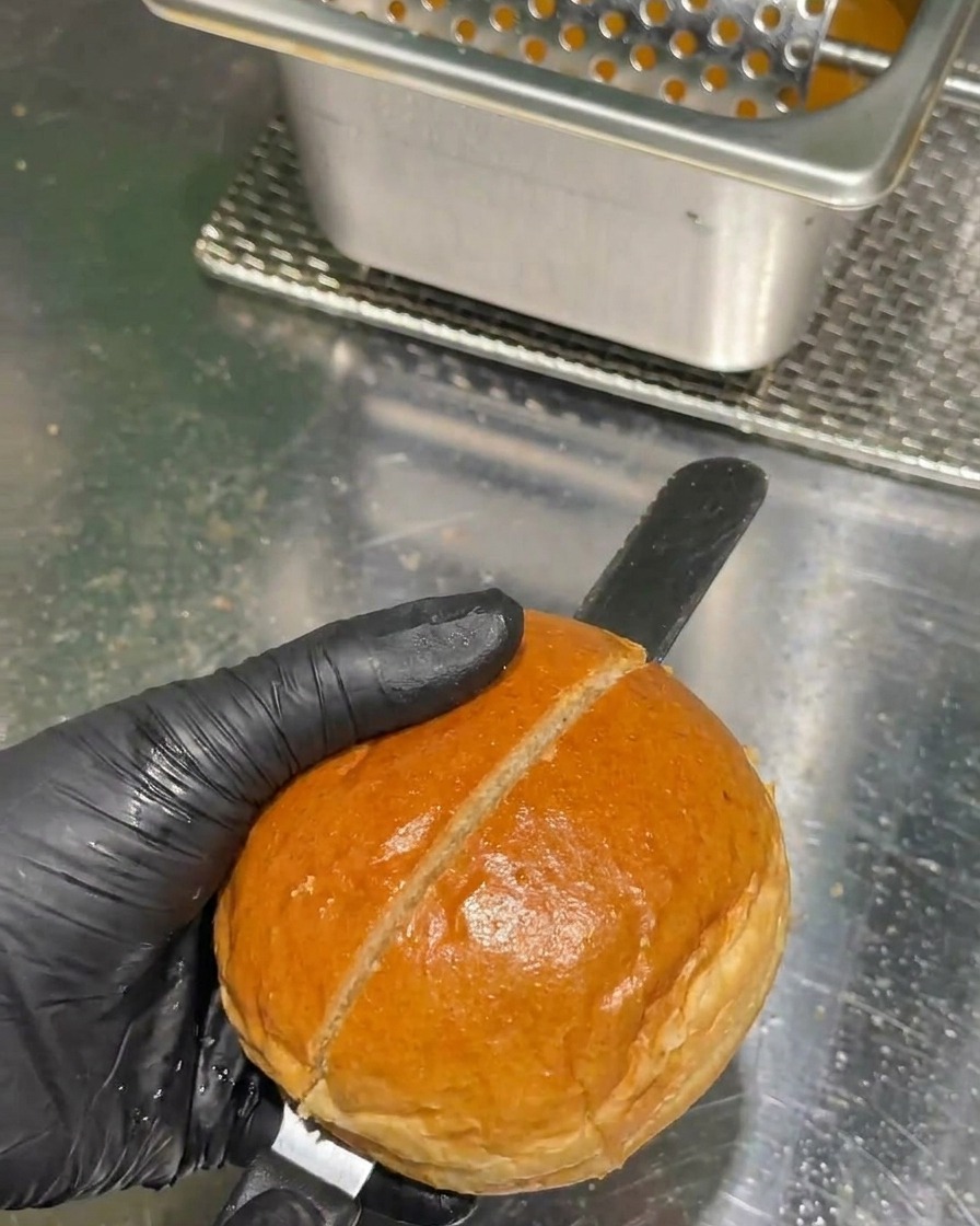 Une main avec un gant noir utilisant un long couteau à pain pour couper un pain à burger doré en deux sur un plan de travail en acier.