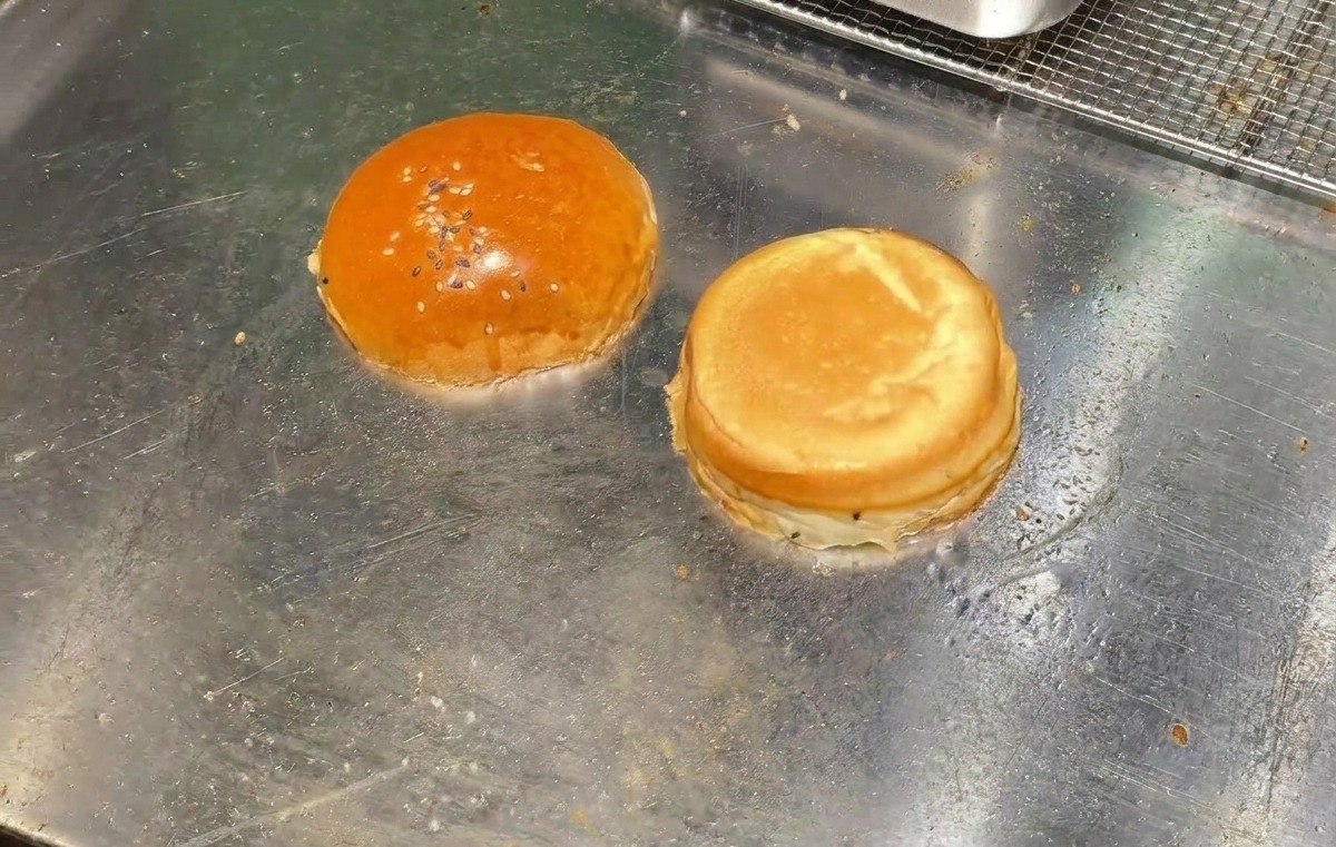 Deux moitiés de pain à burger face coupée vers le bas en train de griller sur une plaque de cuisson en métal.