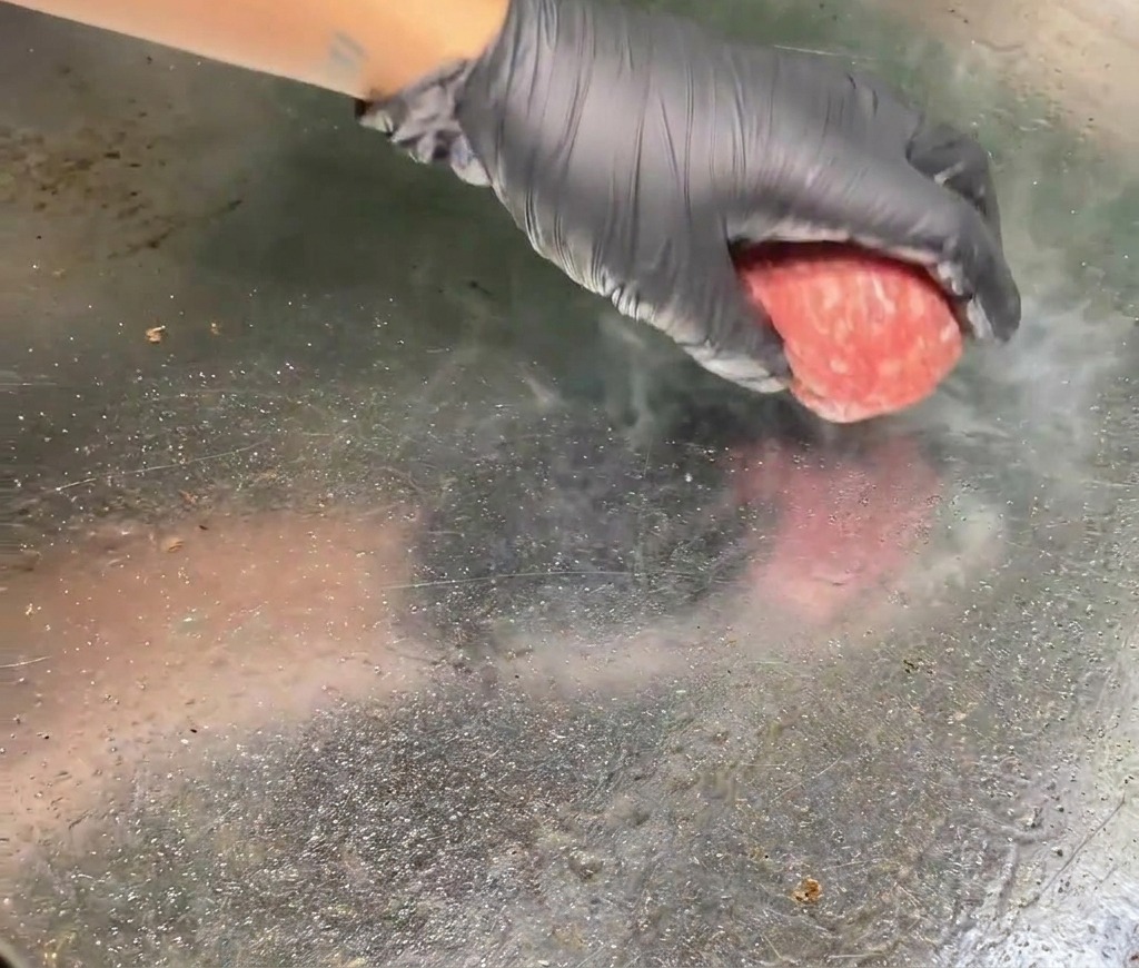 Une main gantée déposant une boule de bœuf haché cru sur un gril brûlant doù séchappe de la vapeur.