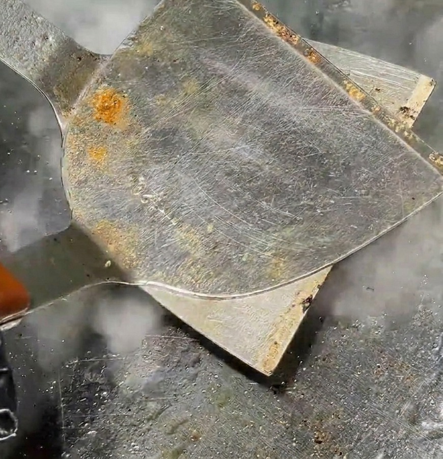 Une spatule métallique écrasant une boule de viande en un steak fin sur un gril chaud avec de la vapeur.