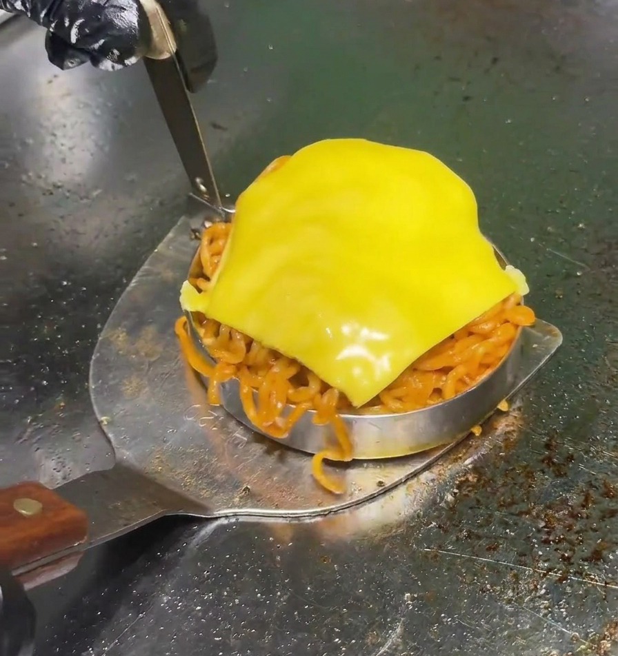 Une spatule métallique soulevant un emporte-pièce rempli de nouilles rouges épicées et surmonté dune tranche de cheddar jaune sur un gril professionnel.