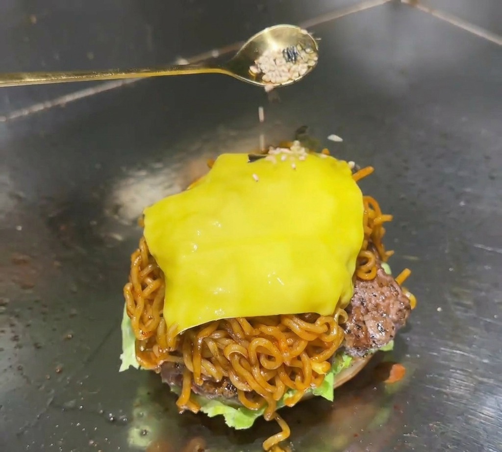 Une cuillère dorée saupoudrant un mélange de graines de sésame blanc et de flocons dalgues noires sur le cheddar fondu dun burger de ramen.