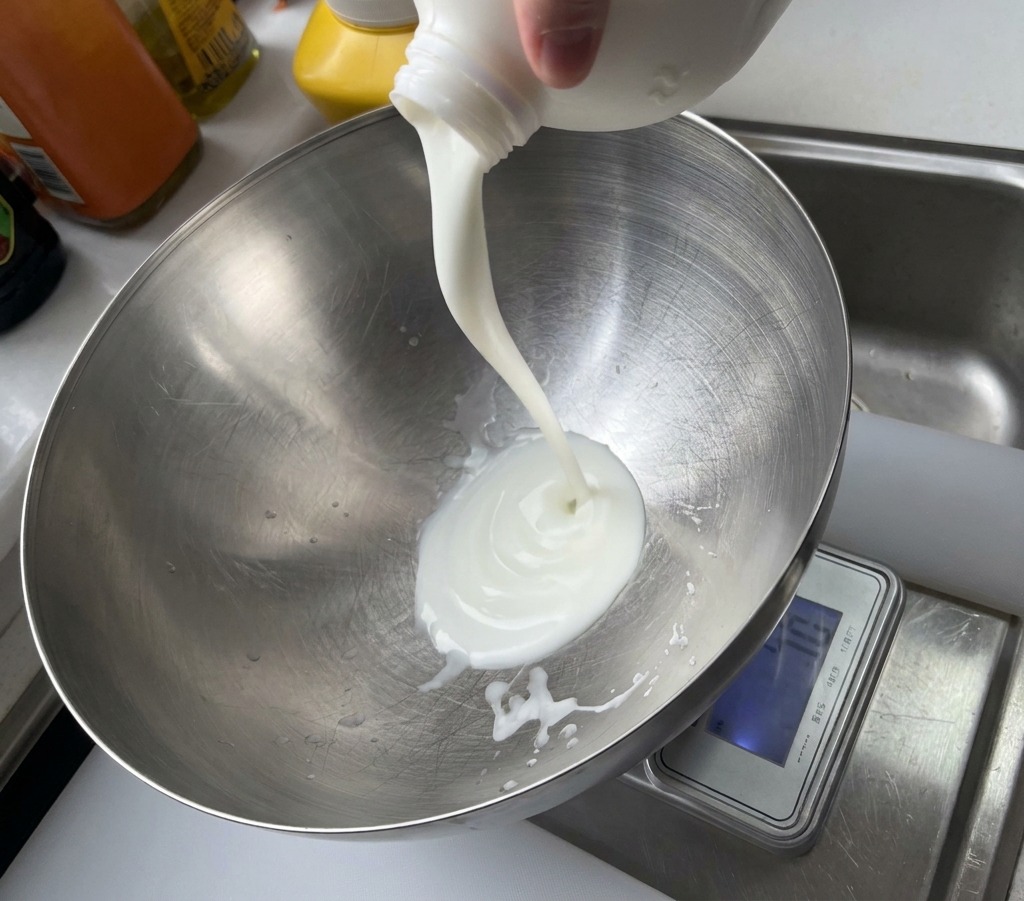 Un filet de yaourt blanc versé depuis un pot en plastique dans un grand saladier en métal.