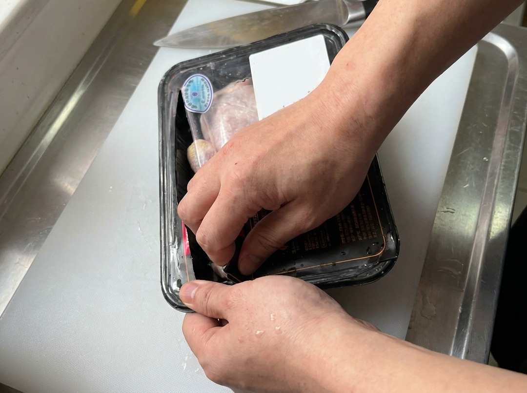 Des mains ouvrant une barquette de hauts de cuisse de poulet crus sur une planche à découper blanche à côté dun couteau de cuisine.