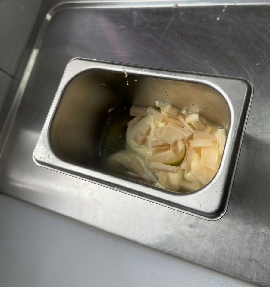 Un récipient en acier inoxydable contenant de la mayonnaise, de la moutarde jaune et des éclats de cornichons hachés.