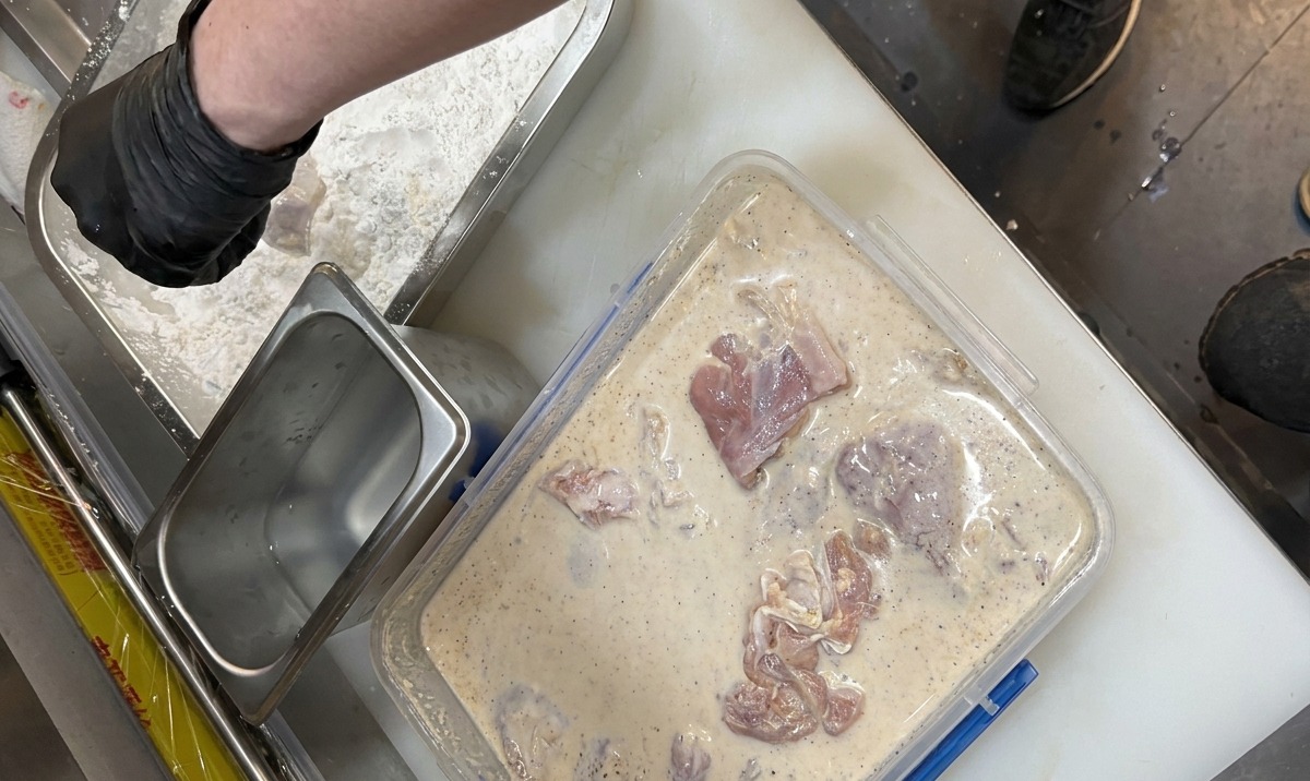 Des mains gantées transférant le poulet mariné dun liquide laiteux vers un plateau de farine denrobage assaisonnée.