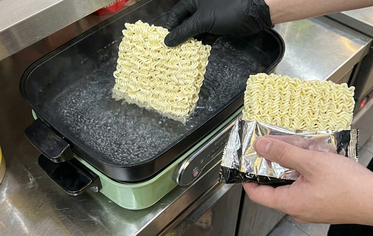 Une personne portant des gants noirs plongeant des blocs carrés de nouilles instantanées sèches dans une casserole deau bouillante.