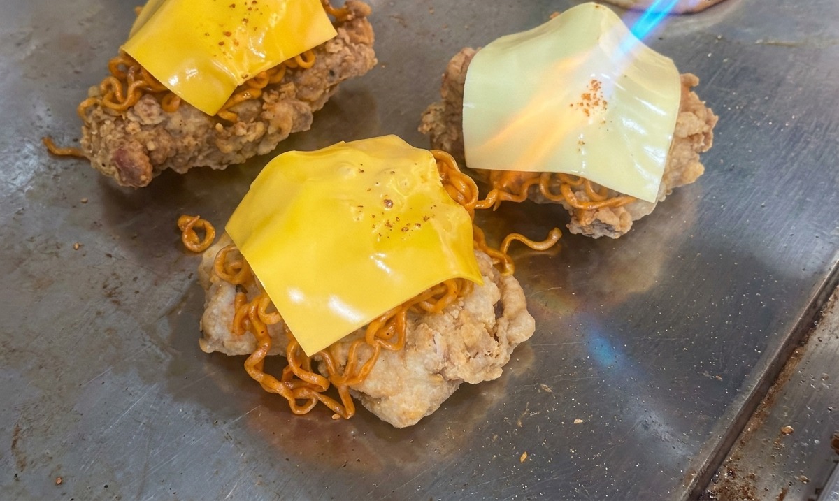 La flamme dun chalumeau faisant fondre une tranche de fromage jaune sur un tas de nouilles épicées et de poulet frit sur un gril.