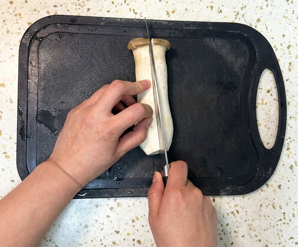 Une personne coupe un gros champignon eryngii en deux dans le sens de la longueur avec un couteau de chef sur une planche à découper noire.