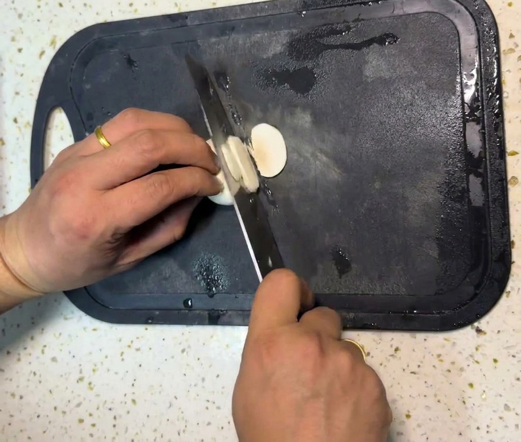 Les mains dune personne éminçant des champignons de Paris blancs en fines rondelles sur une planche à découper noire avec un couteau de chef.