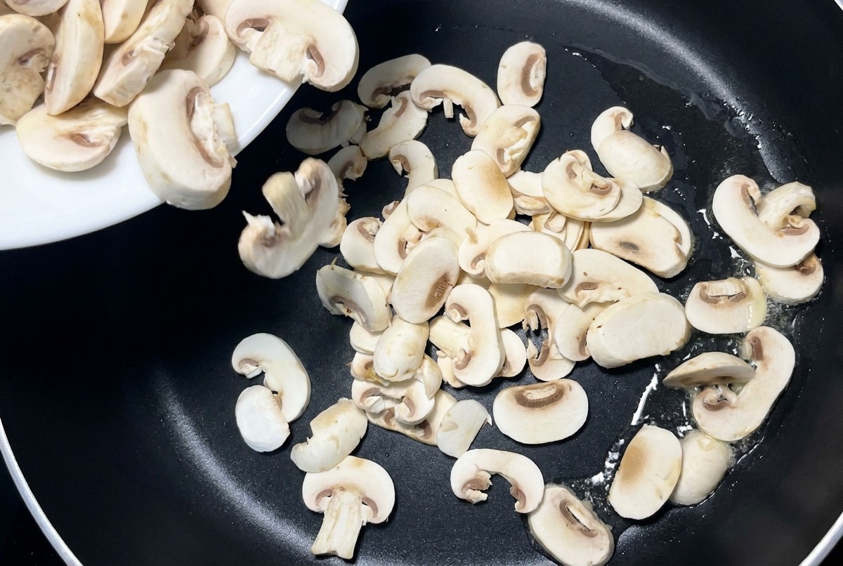 Des lamelles de champignons blancs versées depuis une assiette en céramique blanche dans une poêle antiadhésive noire.