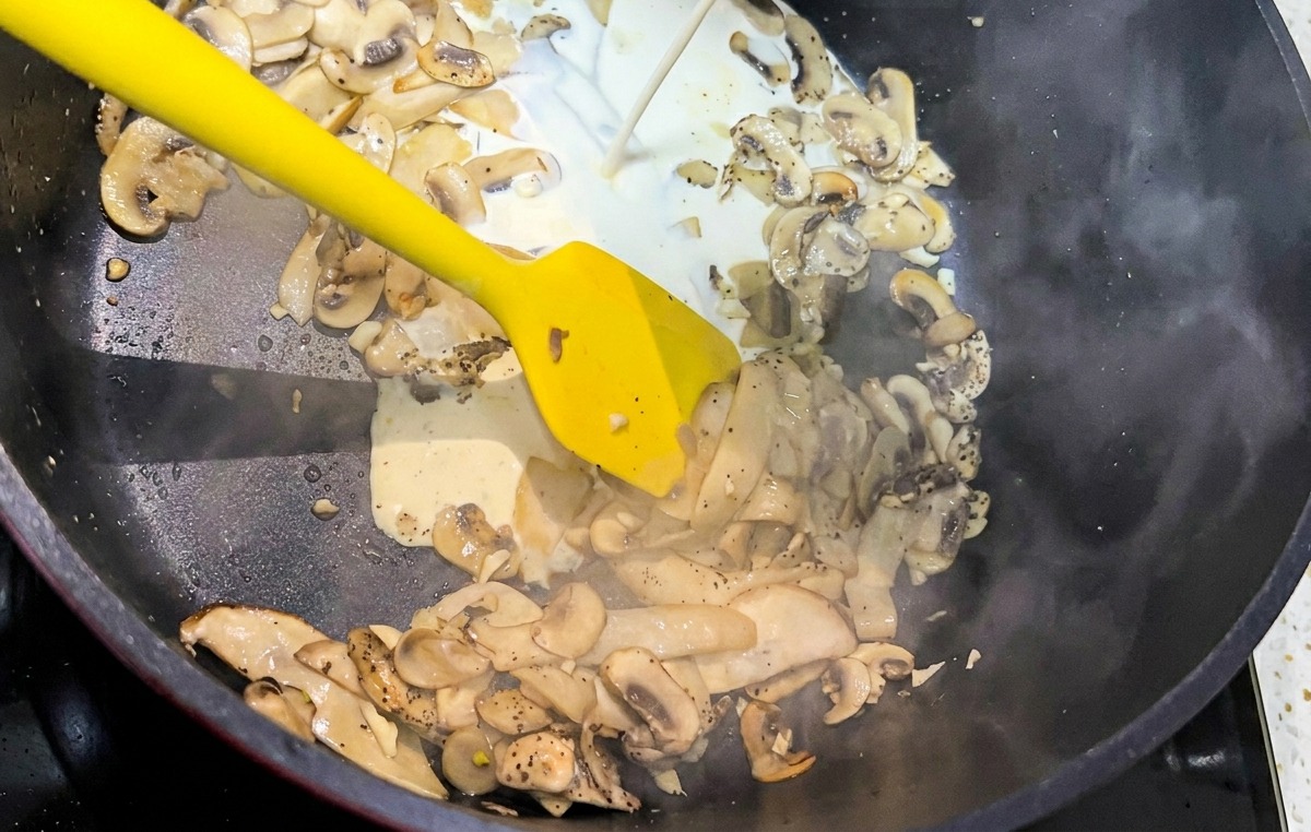 Un filet de crème liquide blanche versé dans une poêle noire remplie de lamelles de champignons sautés.