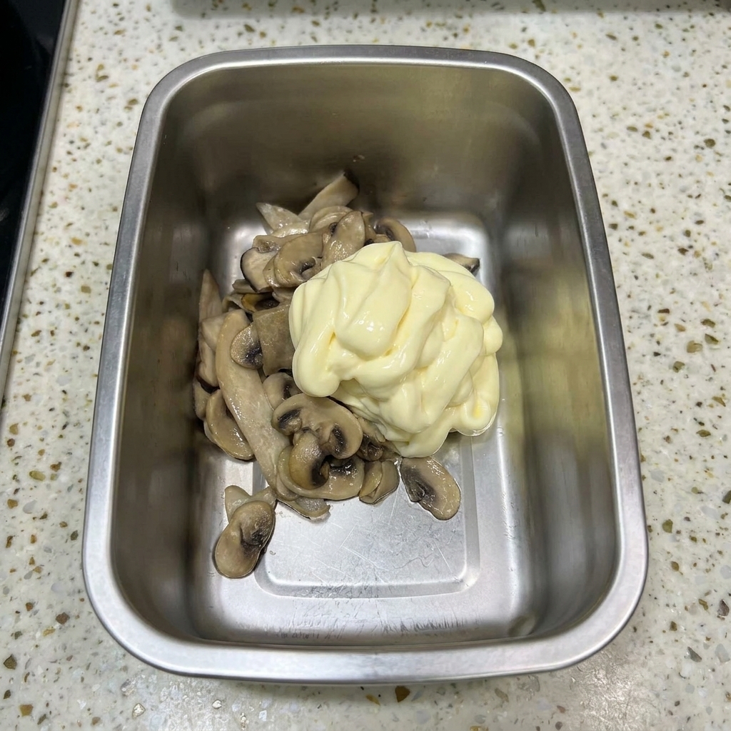 Un récipient en métal contenant les champignons sautés surmontés dun généreux colimaçon de mayonnaise jaune pâle.